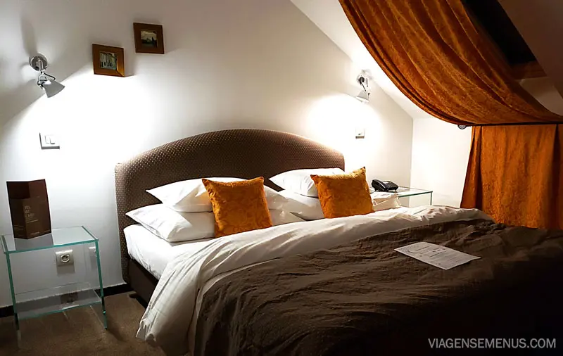 Hotel Leonardo em Praga - quarto de casal, cama king com colcha  e traveseeiros brancos mais dois travesseiros laranja, cortina laranja e 2 abajoures ligados.