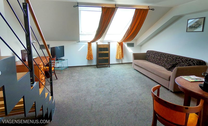 Hotel romântico em Praga - quarto de baixo, com sofá, janela com cortinas laranjas, mesa redonda com cadeira, mesa de escritório, TV e escada