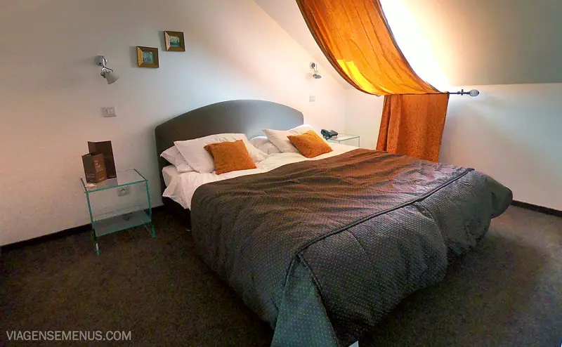 Hotel Leonardo em Praga - quarto de cima, uma cama de casal com 4 travesseiros branco e 2 laranjas, colcha cinza, uma janela com cortina laranja e duas escrivaninhas.