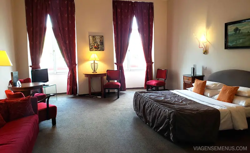 Hotel romântico em Praga - quarto amplo com duas janelas grandes, cortinas grandes até o chão vermelhas, cama King com colcha cinza, duas cadeiras clássicas de estofado vermelho e sofá.