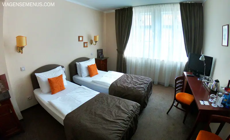 Hotel em Praga - quarto com duas camas de solteiro, colcha cinza escura e travesseiros laranjas, uma janela larga com cortinas verde escuras, escrivaninha com uma cadeira de estofado laranja