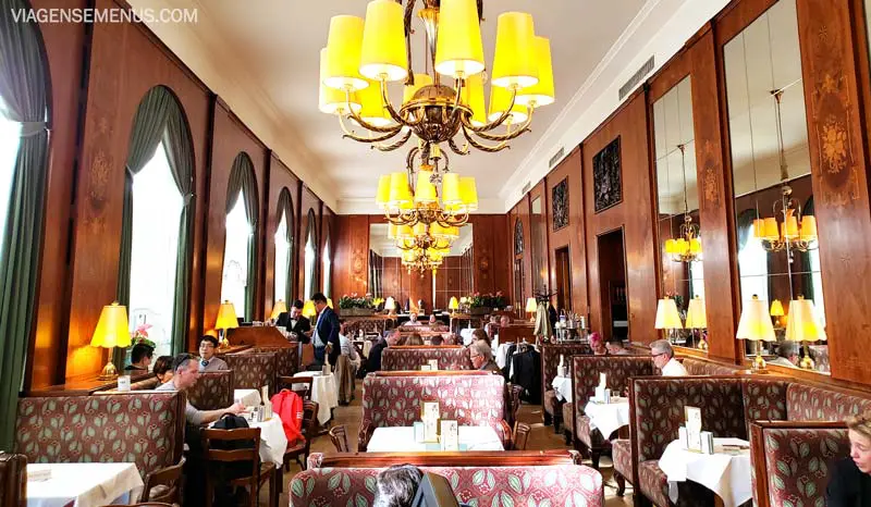 Café em Viena - salão todo no tom marrom, com sofás em estofado marrom com dourado, grandes lustres dourados e paredes com detalhes de madeira